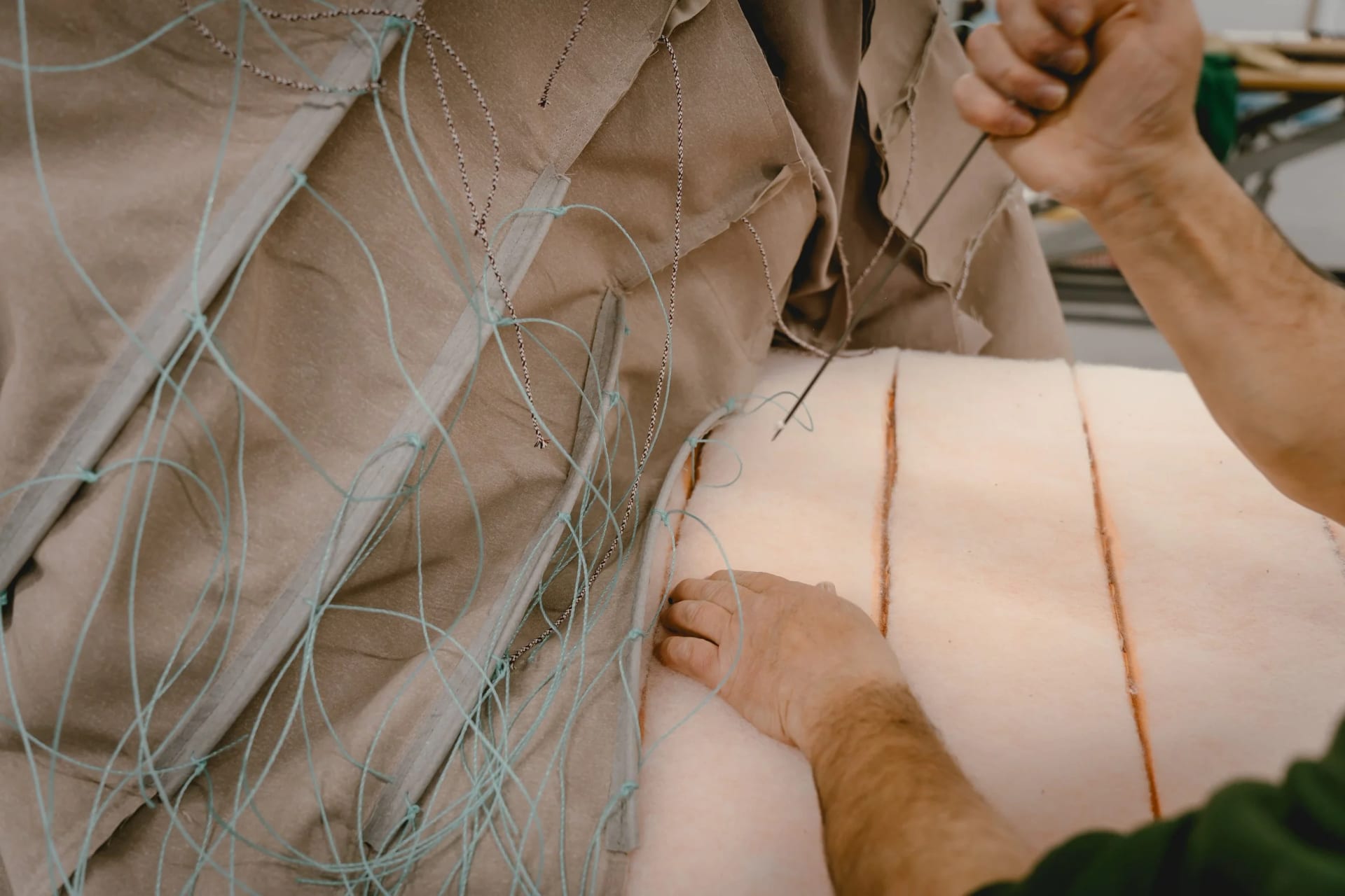 Hands drawing twine through hessian with a long upholstery needle, a roll of wadding alongside