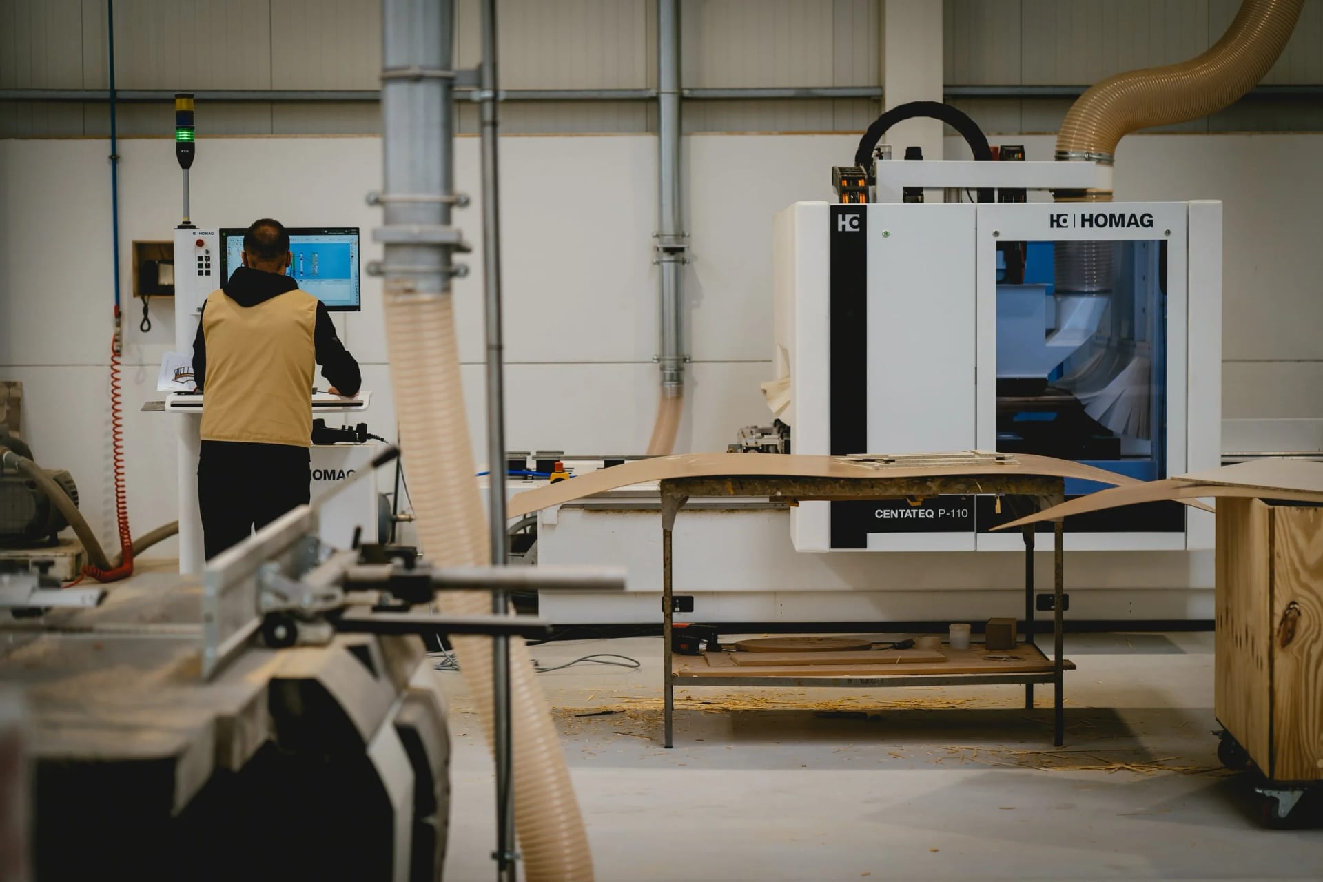 An operator at the console of a large CNC machining center, cut panels stacked on trestles nearby