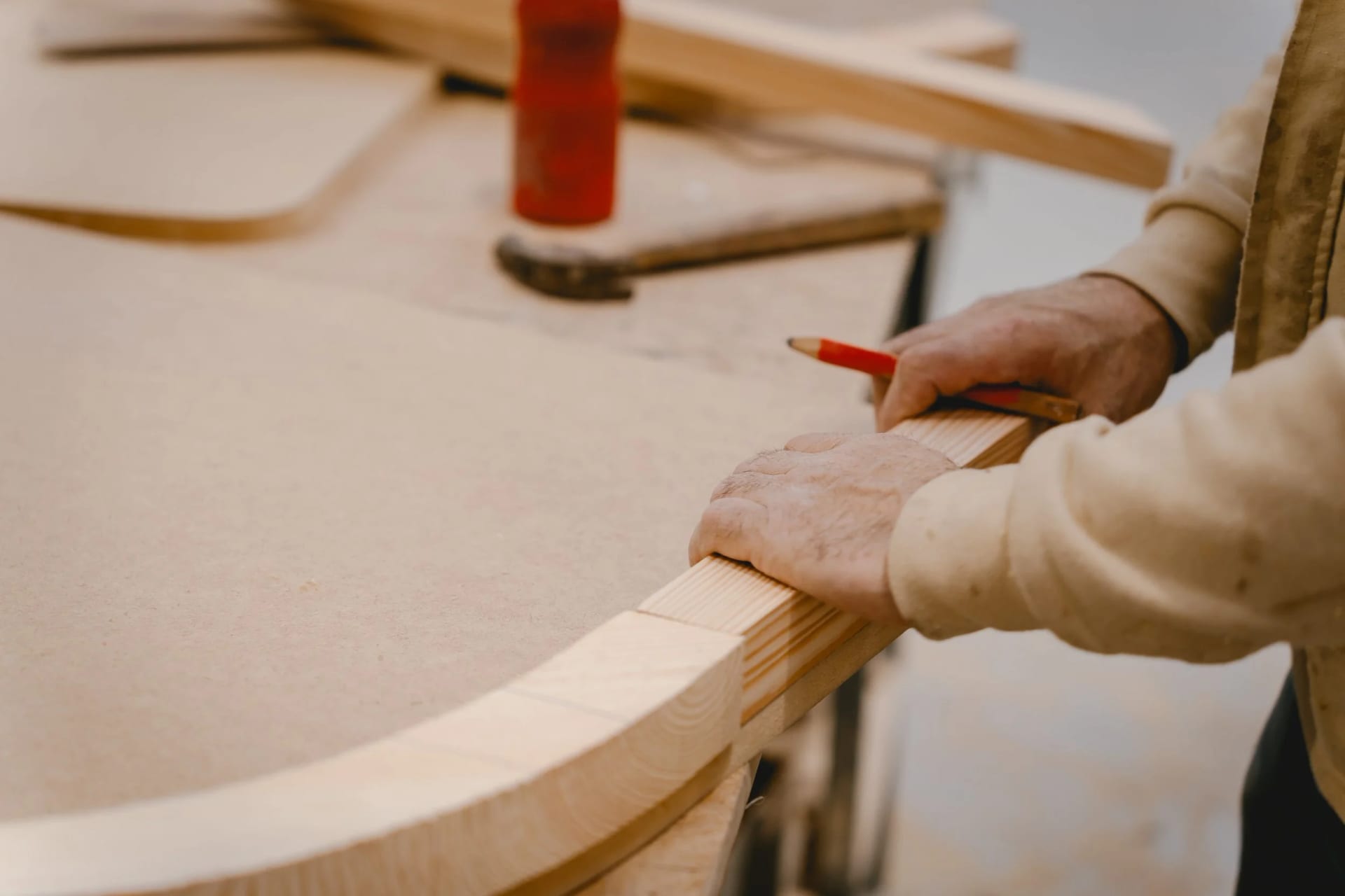 A hand marking a timber section with a carpenter's pencil, a curved laminated frame on the bench beside it