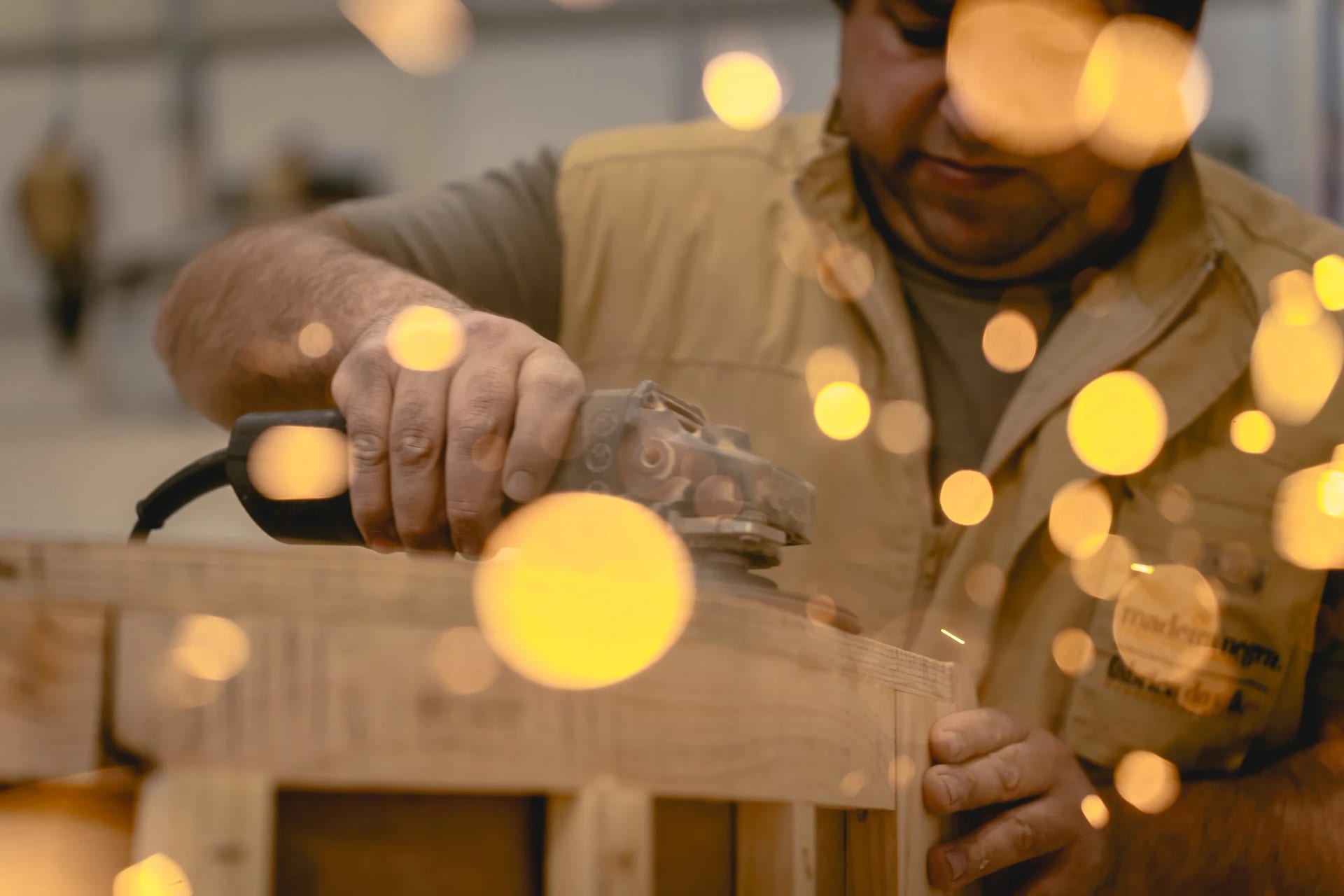 A craftsman working the edge of a wooden frame with a hand sander, warm out-of-focus lights in the foreground